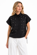 A woman modeling a black short-sleeve button-up shirt with small white embroidered dots, paired with white pants, against a plain white background.