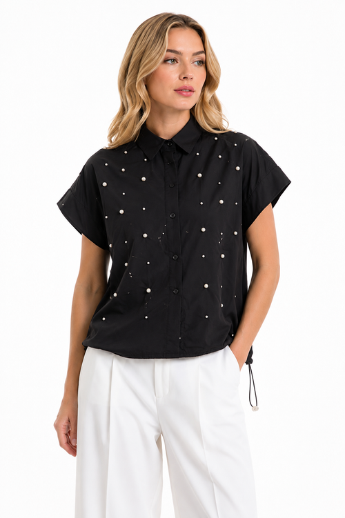 A woman modeling a black short-sleeve button-up shirt with small white embroidered dots, paired with white pants, against a plain white background.