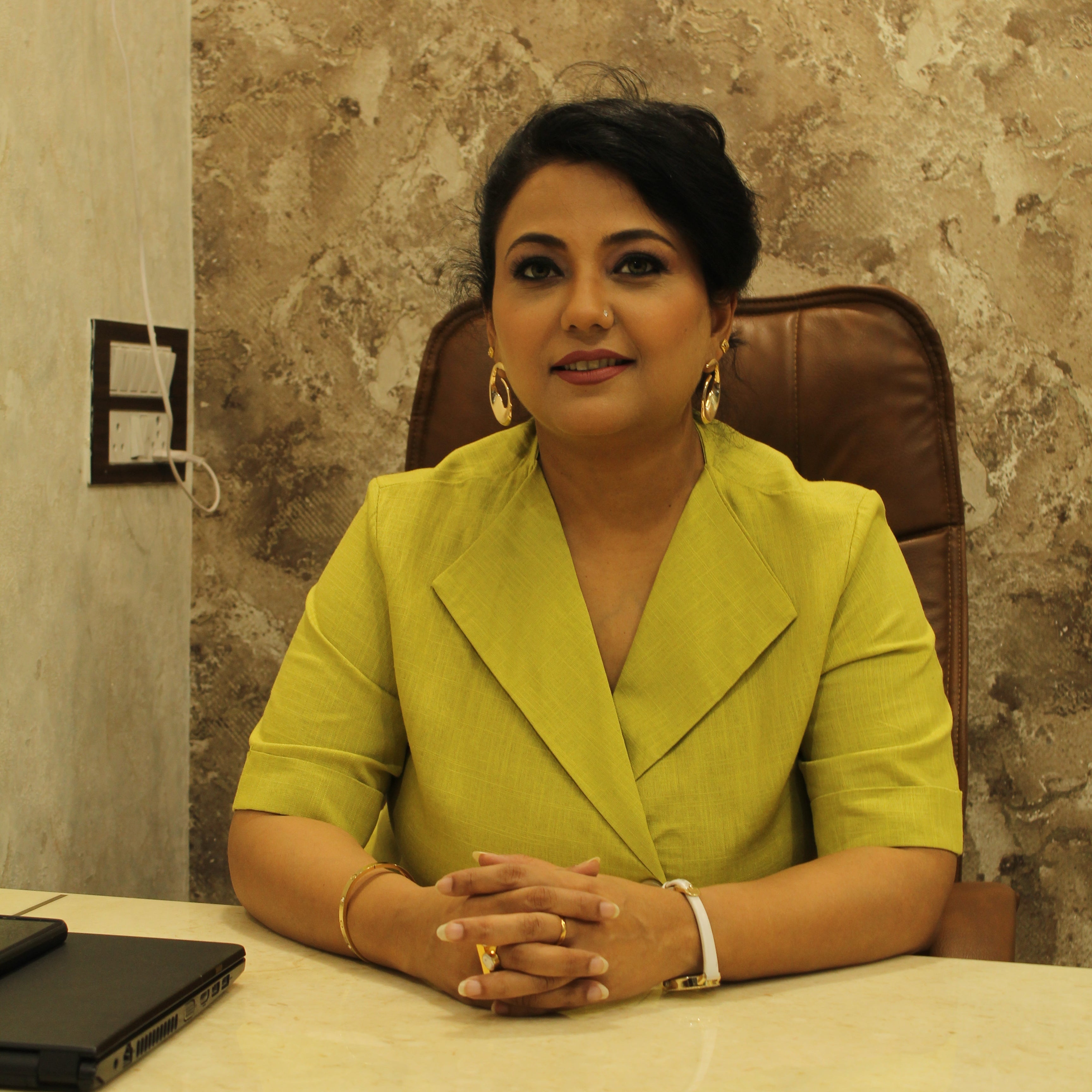 Akanksha Jain in a green blazer sitting at a desk with a decorative wall behind her