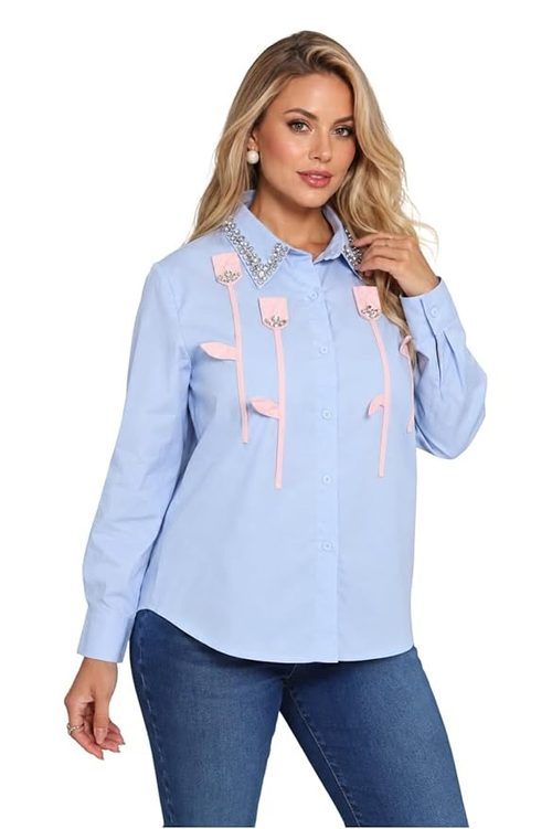 A woman modeling a light blue long-sleeve button-up shirt with pink decorative strap details and silver rhinestone embellishments on the chest, paired with blue jeans against a plain white background.